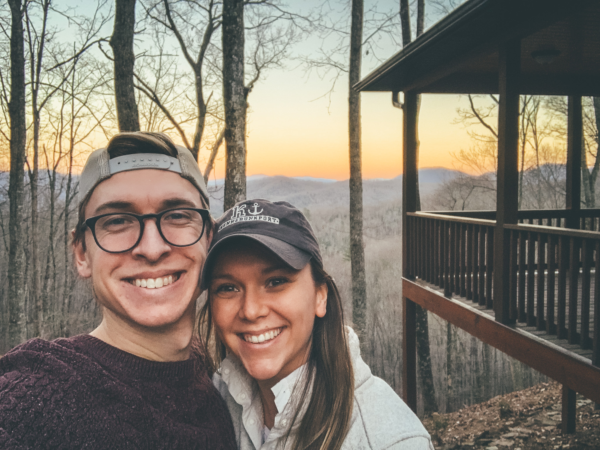 a couple posing in front of a mountain cabin with a sunset in background